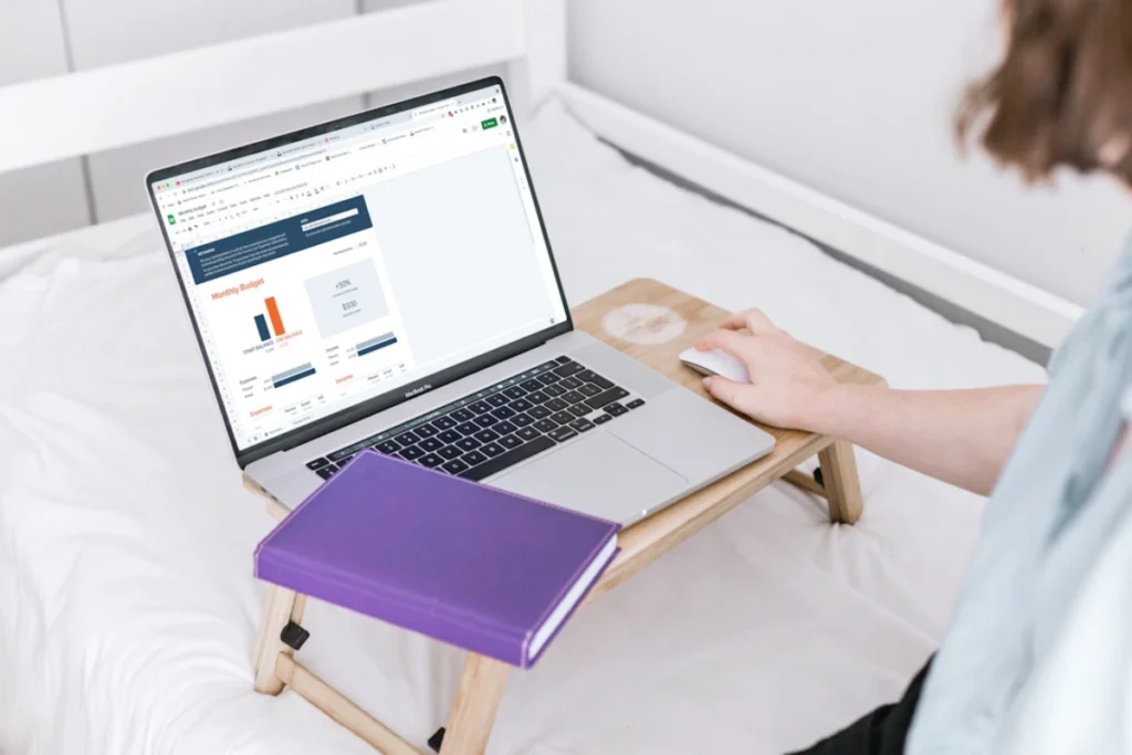 A woman sits on a bed, using a laptop and writing in a notebook, focused on budget and timeline planning.