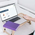A woman sits on a bed, using a laptop and writing in a notebook, focused on budget and timeline planning.