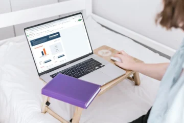 A woman sits on a bed, using a laptop and writing in a notebook, focused on budget and timeline planning.