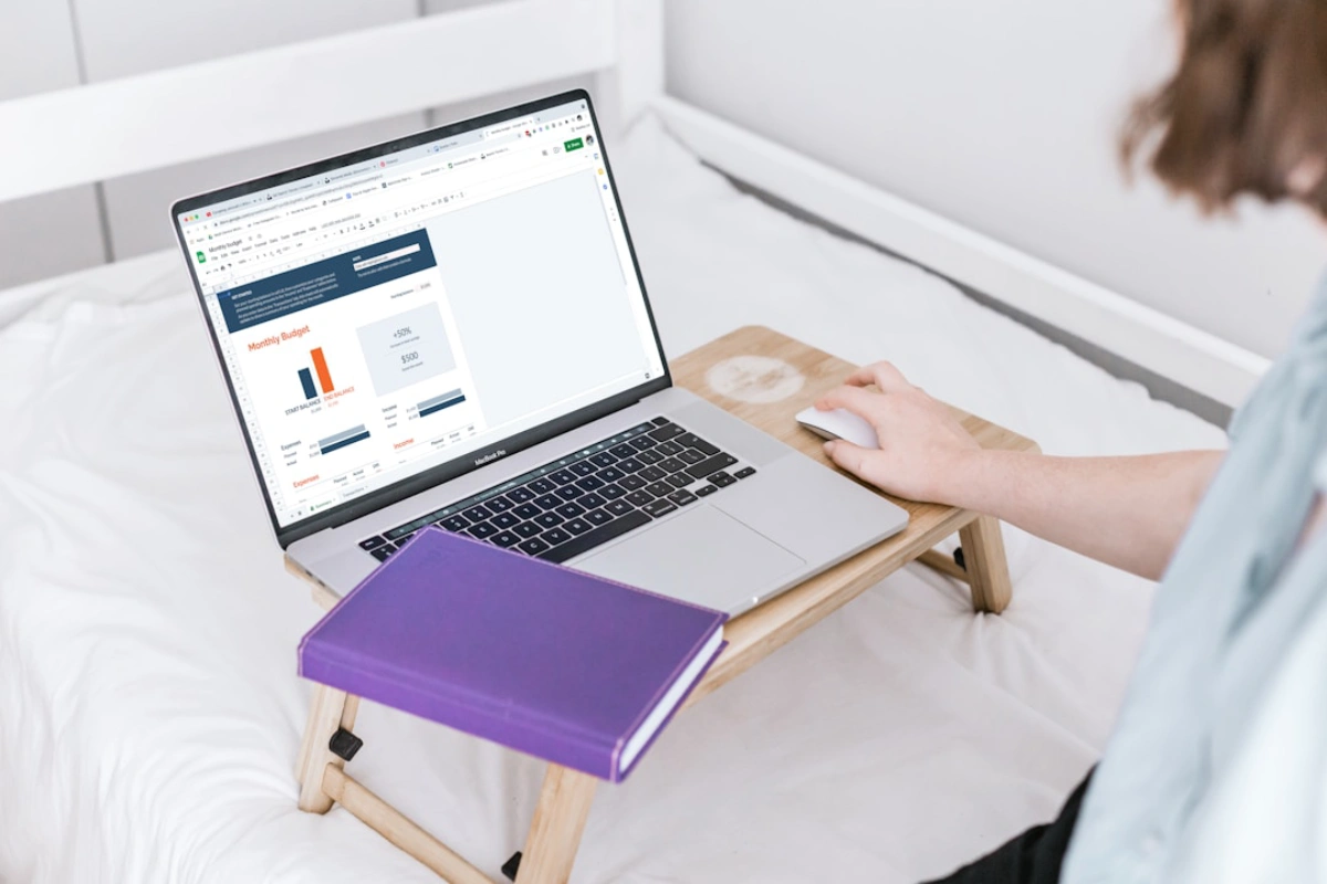 A woman sits on a bed, using a laptop and writing in a notebook, focused on budget and timeline planning.